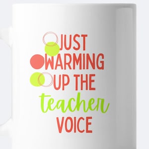 May include: White ceramic mug with the text "Just warming up the teacher voice" in red and green lettering. There are three circles in the design, one red and two green.