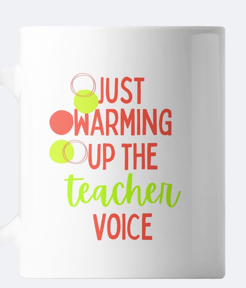 May include: White ceramic mug with the text "Just warming up the teacher voice" in red and green lettering. There are three circles in the design, one red and two green.