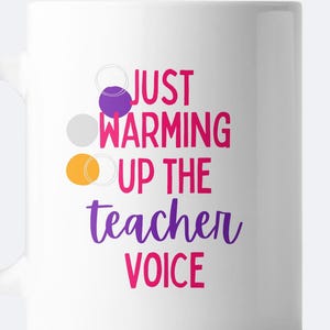 May include: White ceramic mug with the text "Just warming up the teacher voice" in pink and purple lettering. Three colorful circles are arranged vertically on the left side of the mug.