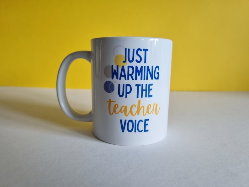 May include: White ceramic mug with a blue and yellow design that says "Just warming up the teacher voice".