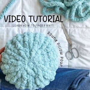 Peut inclure: Un pouf coussin rond tricoté bleu clair. L'image comprend le texte "VIDEO TUTORIAL" et "APPRENDRE À TRICOTER AU DOIGT !" ainsi qu'une paire de ciseaux argentés et un morceau de la même laine. Les mots "Round Pillow Pouf" sont également dans l'image.