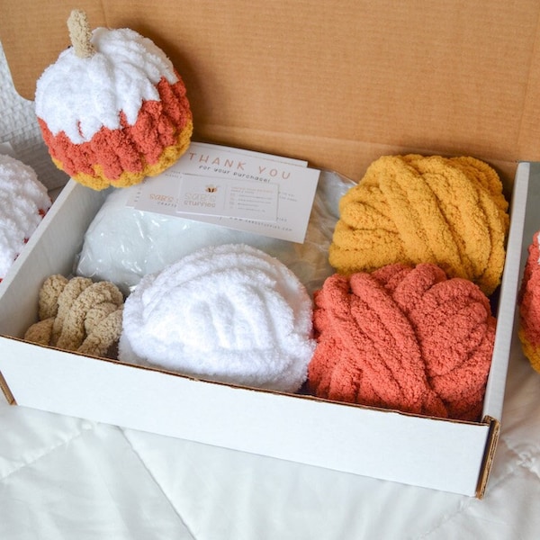 May include: A white box filled with yarn and crocheted pumpkin decorations. The yarn is in shades of orange, yellow, and white. The pumpkins are white with orange and yellow accents. The box also includes a thank you card.