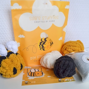 May include: A DIY finger puppet kit with a yellow, black, and white bee theme. The kit includes yarn in white, black, and gray, a bag of stuffing, and instructions. The kit is packaged in a yellow bag with the text "Sab's Stuffies Crafted by Hand" and a bee illustration.