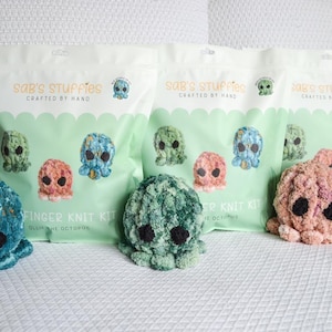 May include: Three finger knit kits for making octopus plushies. The kits are packaged in clear plastic bags with green and white labels that say "SAB's Stuffies Crafted by Hand". The kits include instructions and yarn in teal, green, and pink.
