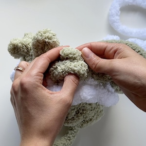 May include: Close-up of hands working with soft, textured yarn in shades of green and white. The yarn appears to be a chunky knit, possibly for a craft project like a blanket or stuffed animal. The focus is on the tactile nature of the materials.