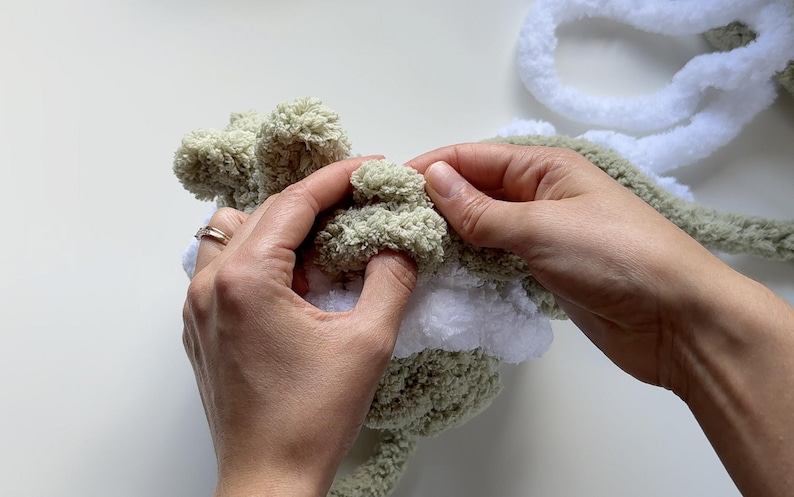May include: Close-up of hands working with soft, textured yarn in shades of green and white. The yarn appears to be a chunky knit, possibly for a craft project like a blanket or stuffed animal. The focus is on the tactile nature of the materials.