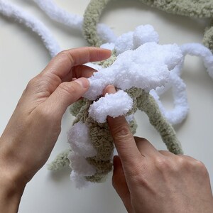 May include: Close-up of hands working with chunky yarn. The yarn is a combination of white and olive green, with a fluffy texture. The hands are manipulating the yarn, possibly for a knitting or crochet project.