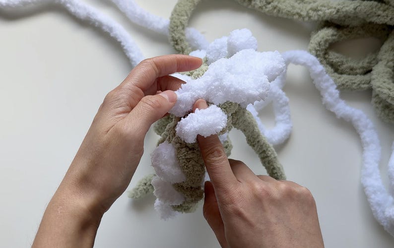May include: Close-up of hands working with chunky yarn. The yarn is a combination of white and olive green, with a fluffy texture. The hands are manipulating the yarn, possibly for a knitting or crochet project.