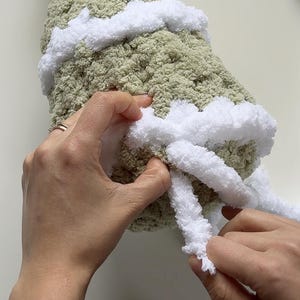 May include: Close-up of hands working with a soft, textured, light green yarn, accented with white yarn. The white yarn is being looped around the green yarn, creating a decorative edge. The craft project is in progress.