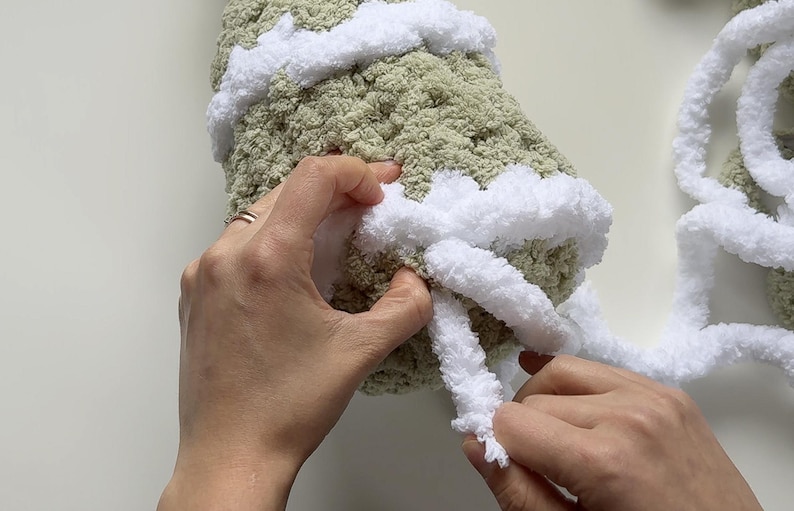 May include: Close-up of hands working with a soft, textured, light green yarn, accented with white yarn. The white yarn is being looped around the green yarn, creating a decorative edge. The craft project is in progress.