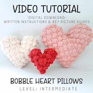 May include: Three heart-shaped pillows in cream, red, and pink. The pillows are made of a textured, chunky yarn. The image includes the text "VIDEO TUTORIAL", "BOBBLE HEART PILLOWS", and "LEVEL: INTERMEDIATE".