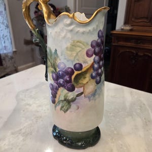 May include: A tall, antique pitcher with a gold-trimmed handle and rim. The body is painted with purple grapes and green leaves. The base is dark green, and the pitcher has a classic, elegant design.