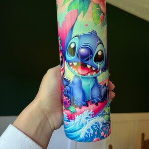 Puede incluir: Un vaso de acero inoxidable azul y rosa con una imagen de dibujos animados de Stitch de Lilo & Stitch montando una ola rosa. El vaso tiene un dise&ntilde;o floral tropical.