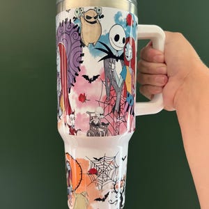 K&ouml;nnte beinhalten: Ein wei&szlig;er Becher mit einem durchsichtigen Deckel und einem gro&szlig;en wei&szlig;en Griff. Der Becher hat ein farbenfrohes Design mit Figuren aus "The Nightmare Before Christmas", darunter Jack Skellington, Sally und Oogie Boogie, mit Flederm&auml;usen und Spinnweben.