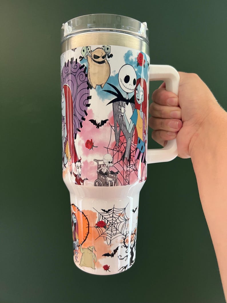 K&ouml;nnte beinhalten: Ein wei&szlig;er Becher mit einem durchsichtigen Deckel und einem gro&szlig;en wei&szlig;en Griff. Der Becher hat ein farbenfrohes Design mit Figuren aus "The Nightmare Before Christmas", darunter Jack Skellington, Sally und Oogie Boogie, mit Flederm&auml;usen und Spinnweben.