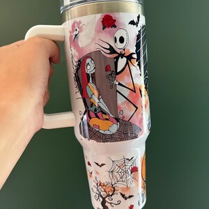 K&ouml;nnte beinhalten: Wei&szlig;er Reisebecher mit Figuren aus The Nightmare Before Christmas. Das Design zeigt Jack Skellington, der Sally eine Rose anbietet, mit Flederm&auml;usen, Spinnweben und einem gruseligen Baum. Der Becher hat einen wei&szlig;en Griff und einen transparenten Deckel.