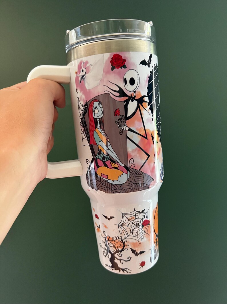 K&ouml;nnte beinhalten: Wei&szlig;er Reisebecher mit Figuren aus The Nightmare Before Christmas. Das Design zeigt Jack Skellington, der Sally eine Rose anbietet, mit Flederm&auml;usen, Spinnweben und einem gruseligen Baum. Der Becher hat einen wei&szlig;en Griff und einen transparenten Deckel.