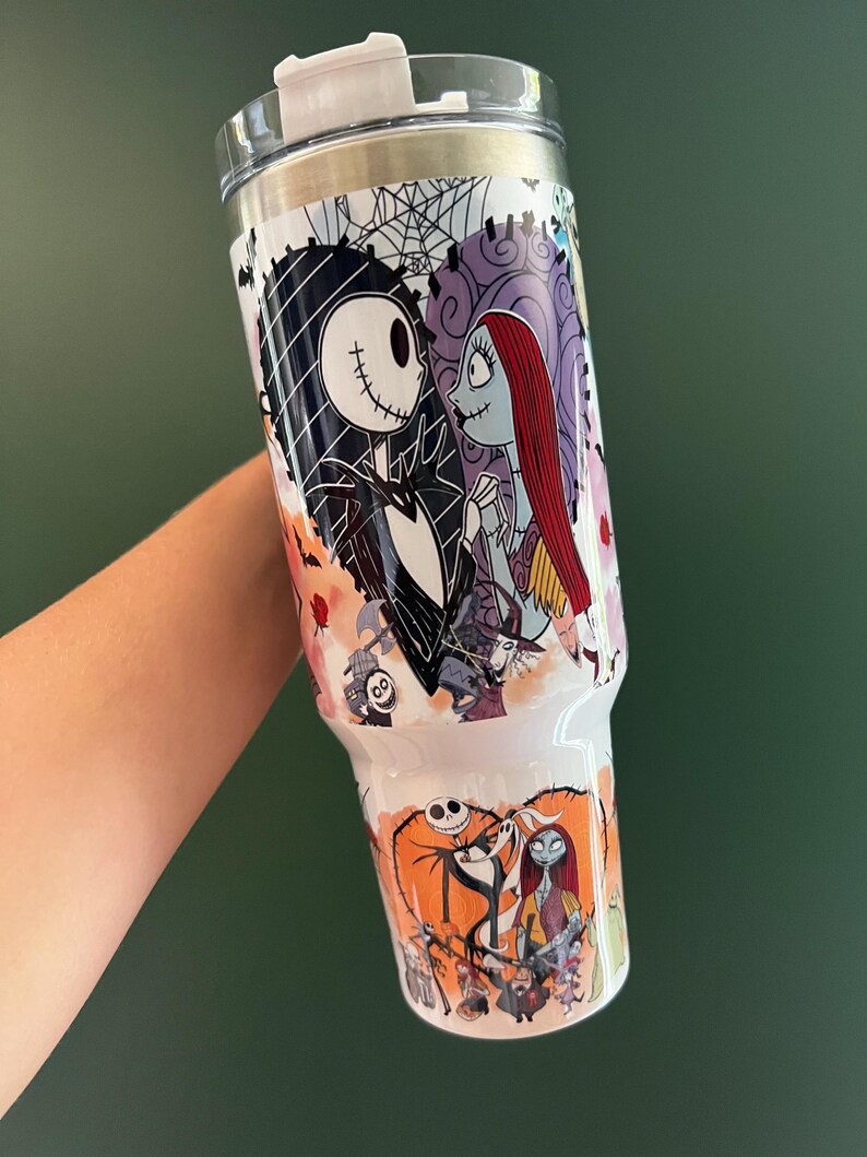K&ouml;nnte beinhalten: Ein wei&szlig;er Becher mit Jack Skellington und Sally aus The Nightmare Before Christmas. Das Design umfasst einen herzf&ouml;rmigen Hintergrund mit einem Spinnennetz, Rosen und anderen Charakteren. Der Becher hat einen wei&szlig;en Deckel und einen silbernen Rand.