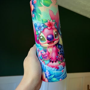Puede incluir: Un vaso de acero inoxidable rosa y azul con una imagen de dibujos animados de Stitch y Angel de Lilo & Stitch. Los personajes llevan collares de flores y est&aacute;n montando una ola. El fondo es una escena tropical con palmeras y flores.