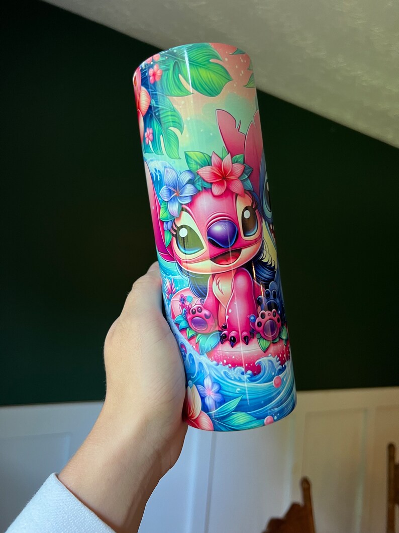 Puede incluir: Un vaso de acero inoxidable rosa y azul con una imagen de dibujos animados de Stitch y Angel de Lilo & Stitch. Los personajes llevan collares de flores y est&aacute;n montando una ola. El fondo es una escena tropical con palmeras y flores.