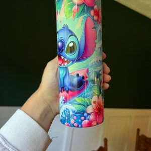 Puede incluir: Un vaso de acero inoxidable azul y rosa con una imagen de dibujos animados de Stitch de Lilo & Stitch haciendo surf en una ola. El vaso est&aacute; rodeado de flores y hojas tropicales.