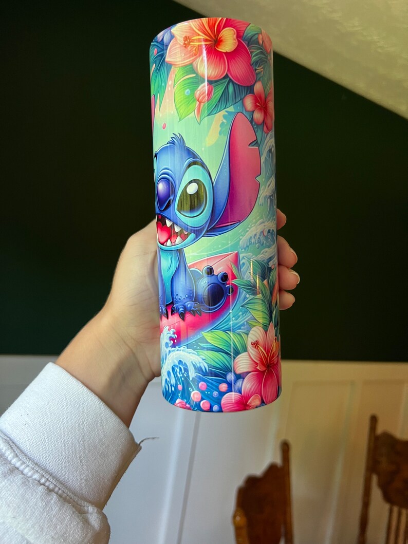 Puede incluir: Un vaso de acero inoxidable azul y rosa con una imagen de dibujos animados de Stitch de Lilo & Stitch haciendo surf en una ola. El vaso est&aacute; rodeado de flores y hojas tropicales.