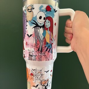 K&ouml;nnte beinhalten: Ein wei&szlig;er Isolierbecher mit Figuren aus The Nightmare Before Christmas. Das Design umfasst Jack Skellington, Sally und andere gruselige Elemente. Der Becher hat einen wei&szlig;en Griff und einen transparenten Deckel.