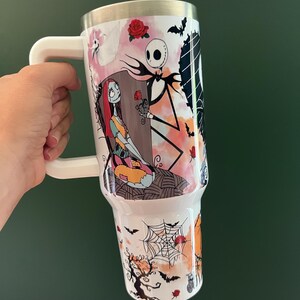K&ouml;nnte beinhalten: Wei&szlig;er Becher mit Henkel und farbenfrohem Design aus The Nightmare Before Christmas. Das Design zeigt Jack Skellington, Sally, Flederm&auml;use, Rosen und ein Spinnennetz. Der Becher hat einen silbernen Rand und einen wei&szlig;en Griff.