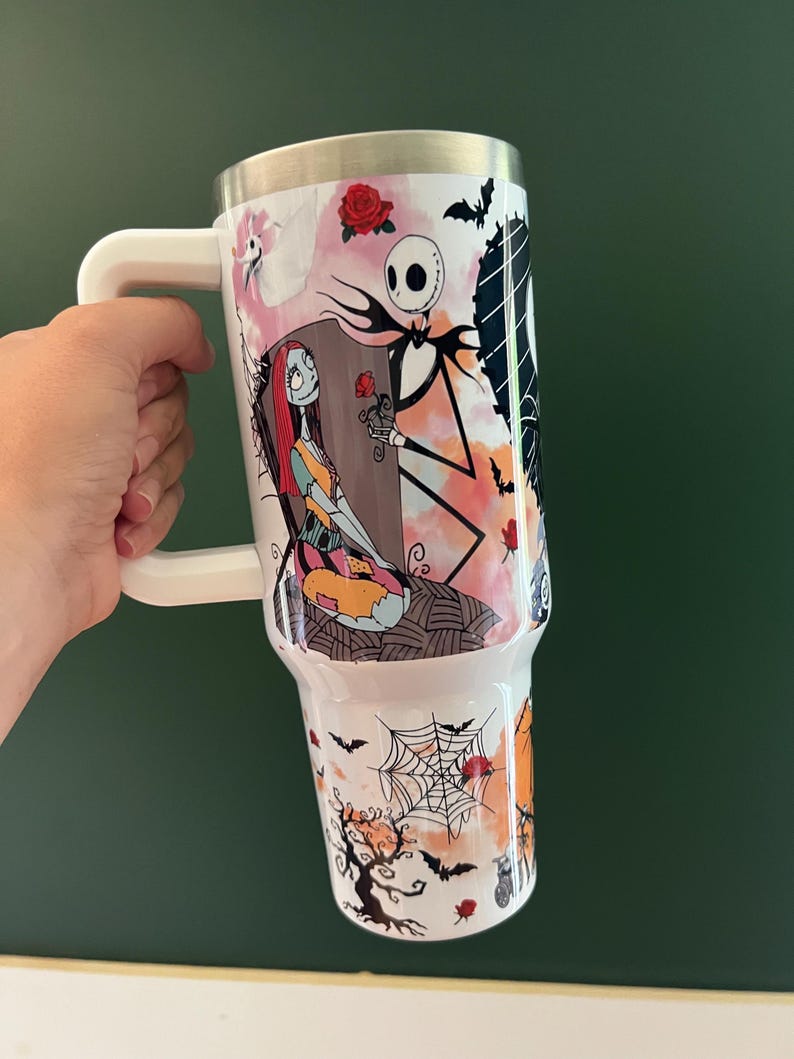 K&ouml;nnte beinhalten: Wei&szlig;er Becher mit Henkel und farbenfrohem Design aus The Nightmare Before Christmas. Das Design zeigt Jack Skellington, Sally, Flederm&auml;use, Rosen und ein Spinnennetz. Der Becher hat einen silbernen Rand und einen wei&szlig;en Griff.