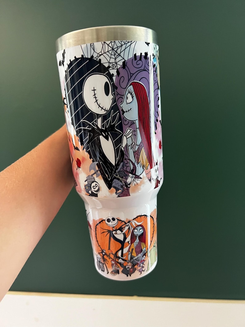 K&ouml;nnte beinhalten: Ein wei&szlig;er Becher mit Illustrationen von Jack Skellington und Sally aus The Nightmare Before Christmas. Das Design umfasst Bilder von Jack und Sally, ein Spinnennetz und ein orangefarbenes Herz. Der Becher hat einen silbernen Deckel.