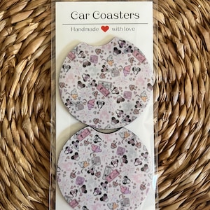 May include: Two round car coasters with a pink and white pattern featuring cartoon mice, hearts, and other whimsical designs. The coasters are packaged in a clear plastic bag with a white card that reads "Car Coasters Handmade with love".