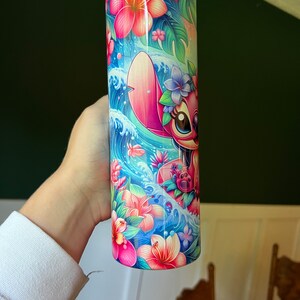 Puede incluir: Un vaso de acero inoxidable rosa y azul con un dise&ntilde;o de personaje de dibujos animados. El personaje es un extraterrestre rosa con ojos azules y una corona de flores. El fondo presenta olas azules, flores rosas y hojas verdes.