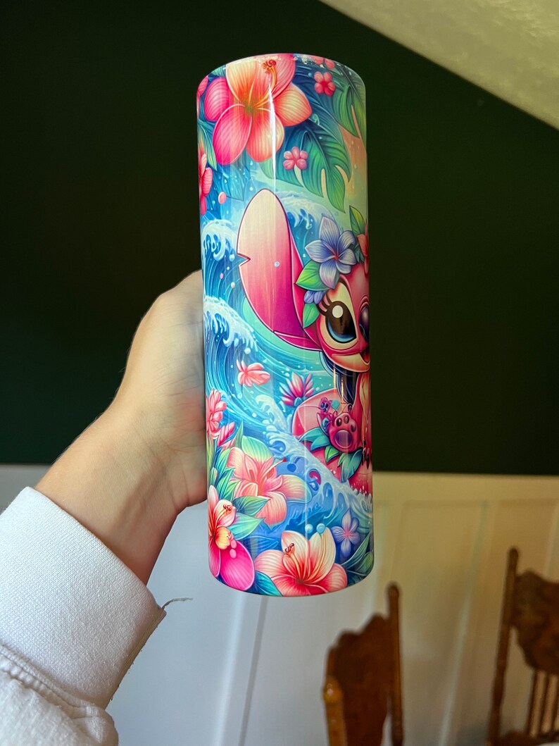 Puede incluir: Un vaso de acero inoxidable rosa y azul con un dise&ntilde;o de personaje de dibujos animados. El personaje es un extraterrestre rosa con ojos azules y una corona de flores. El fondo presenta olas azules, flores rosas y hojas verdes.