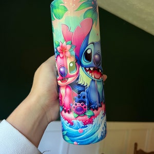 Puede incluir: Un vaso de acero inoxidable azul y rosa con una imagen de dibujos animados de Stitch y Angel de la pel&iacute;cula de Disney Lilo & Stitch. El vaso tiene un tema tropical con hojas de palma y flores.