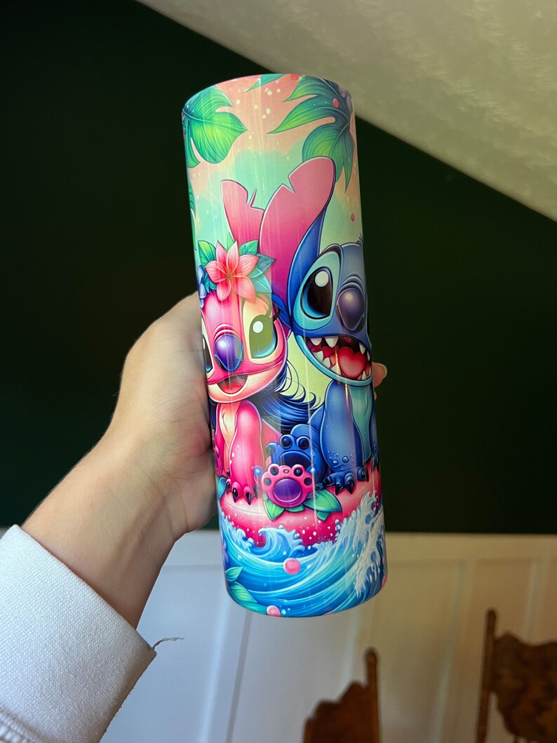 Puede incluir: Un vaso de acero inoxidable azul y rosa con una imagen de dibujos animados de Stitch y Angel de la pel&iacute;cula de Disney Lilo & Stitch. El vaso tiene un tema tropical con hojas de palma y flores.