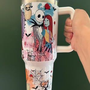 K&ouml;nnte beinhalten: Wei&szlig;er Reisebecher mit Jack Skellington und Sally aus The Nightmare Before Christmas. Der Becher hat einen wei&szlig;en Griff und Deckel und ist mit Flederm&auml;usen, Rosen und Spinnweben verziert. Der Becher ist etwa 23 cm hoch.