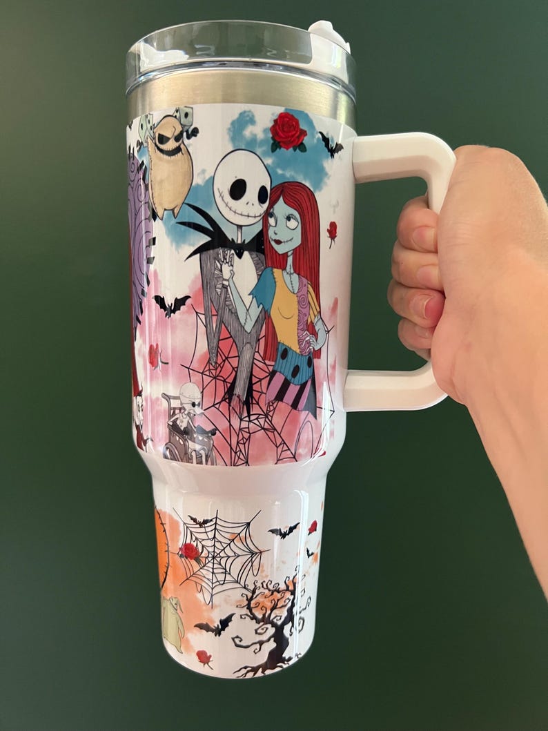 K&ouml;nnte beinhalten: Wei&szlig;er Reisebecher mit Jack Skellington und Sally aus The Nightmare Before Christmas. Der Becher hat einen wei&szlig;en Griff und Deckel und ist mit Flederm&auml;usen, Rosen und Spinnweben verziert. Der Becher ist etwa 23 cm hoch.