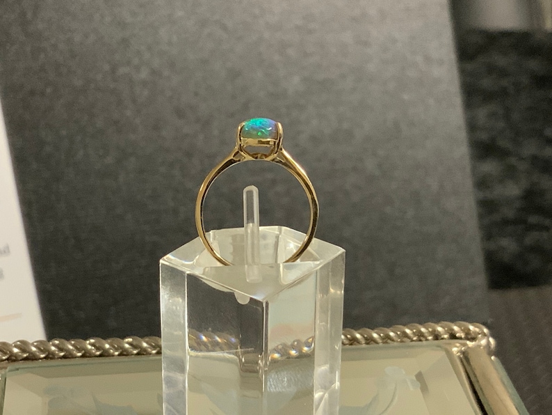 May include: A gold ring with a green opal gemstone. The ring is on a clear display stand.