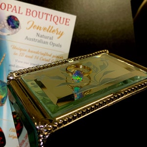May include: A gold ring with a round, iridescent opal gemstone. The ring is displayed in a mirrored jewelry box with a decorative border.