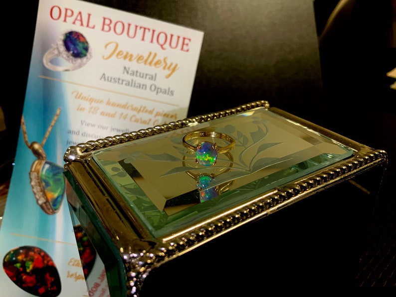 May include: A gold ring with a round, iridescent opal gemstone. The ring is displayed in a mirrored jewelry box with a decorative border.