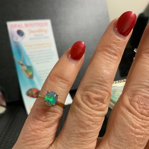 May include: A gold ring with a green, blue, and black opal gemstone. The ring is on a person's finger.