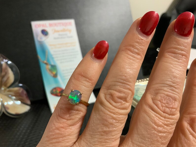 May include: A gold ring with a green, blue, and black opal gemstone. The ring is on a person's finger.