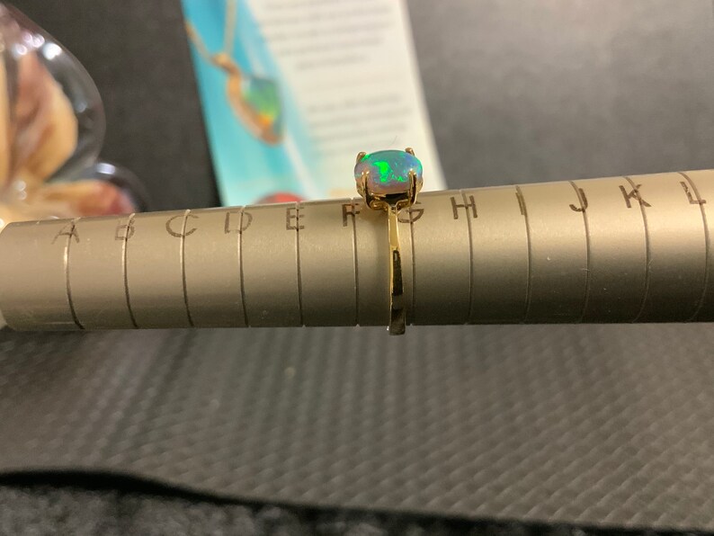 May include: A gold ring with a green and white opal gemstone. The ring is sized on a ring sizer with letters A through L.