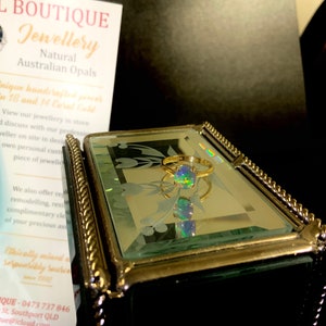 May include: A gold ring with a green, blue, and black opal gemstone. The ring is sitting in a mirrored jewelry box with a gold trim.