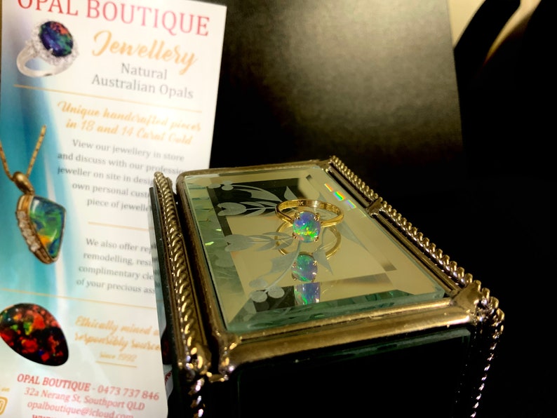 May include: A gold ring with a green, blue, and black opal gemstone. The ring is sitting in a mirrored jewelry box with a gold trim.