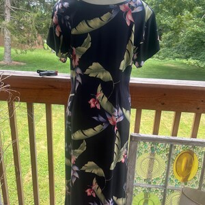 K&ouml;nnte beinhalten: Ein schwarzes Maxikleid mit tropischem Blumendruck. Das Kleid hat kurze &Auml;rmel und einen Rundhalsausschnitt. Der Druck umfasst gro&szlig;e gr&uuml;ne Bl&auml;tter und rosa und rote Blumen. Das Kleid wird auf einer Schaufensterpuppe auf einer Holzterrasse pr&auml;sentiert.