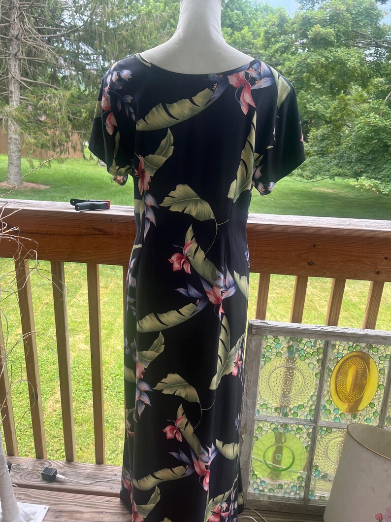 K&ouml;nnte beinhalten: Ein schwarzes Maxikleid mit tropischem Blumendruck. Das Kleid hat kurze &Auml;rmel und einen Rundhalsausschnitt. Der Druck umfasst gro&szlig;e gr&uuml;ne Bl&auml;tter und rosa und rote Blumen. Das Kleid wird auf einer Schaufensterpuppe auf einer Holzterrasse pr&auml;sentiert.