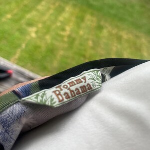 K&ouml;nnte beinhalten: Nahaufnahme eines Tommy Bahama-Etiketts auf einem gemusterten Hemd. Das Etikett ist rechteckig mit dem Markennamen in einer Serifenschrift. Das Hemd hat ein Karomuster mit gr&uuml;nen, blauen und schwarzen Farben.