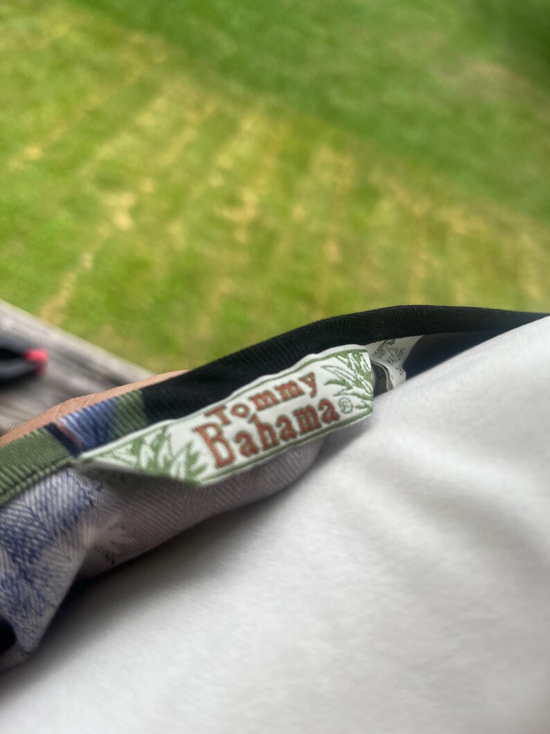 K&ouml;nnte beinhalten: Nahaufnahme eines Tommy Bahama-Etiketts auf einem gemusterten Hemd. Das Etikett ist rechteckig mit dem Markennamen in einer Serifenschrift. Das Hemd hat ein Karomuster mit gr&uuml;nen, blauen und schwarzen Farben.