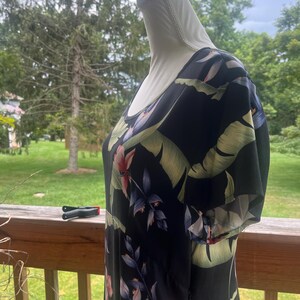 K&ouml;nnte beinhalten: Schwarzes Kleid mit tropischem Blumendruck mit rosa, blauen und gr&uuml;nen Blumen und Bl&auml;ttern. Das Kleid hat kurze &Auml;rmel und einen Rundhalsausschnitt. Das Kleid wird auf einer Schaufensterpuppe pr&auml;sentiert.